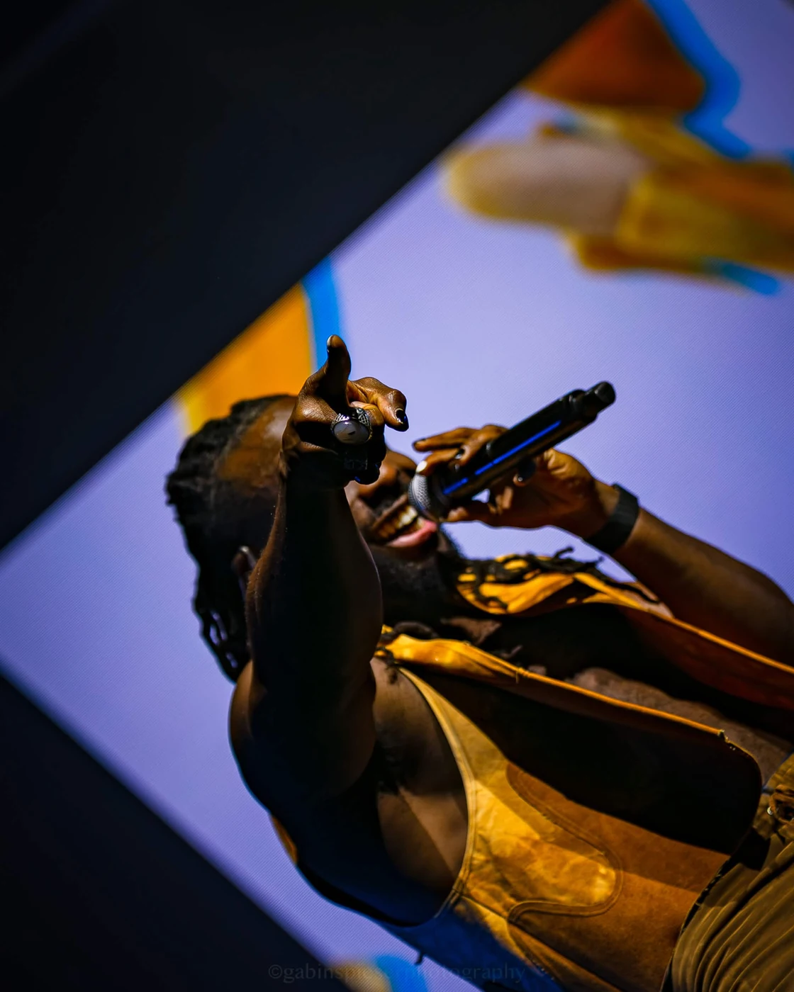 Chanteur · Electro Symphony — En contre-plongée, un chanteur en gilet jaune pointe la main vers l’objectif, micro aux lèvres. Reims Arena · 12 avril 2026.