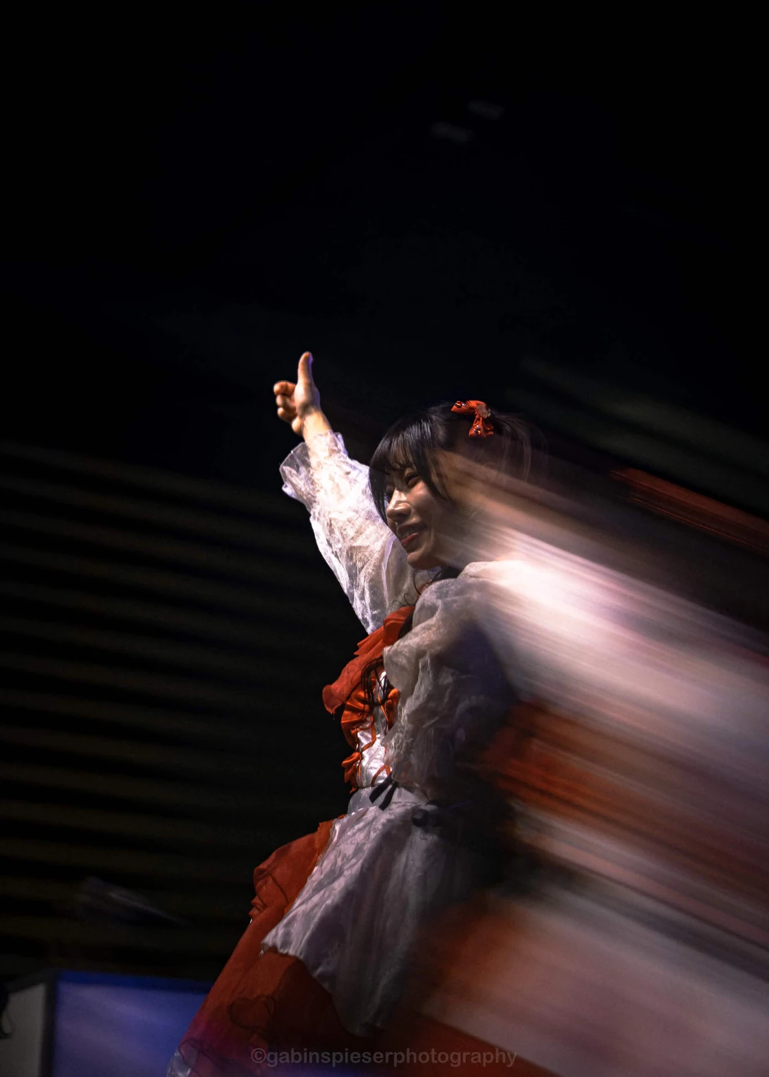 dela — Une artiste en rouge et blanc lève le bras, entourée de traînées de mouvement. Japan Mania · Reims · mai 2026.