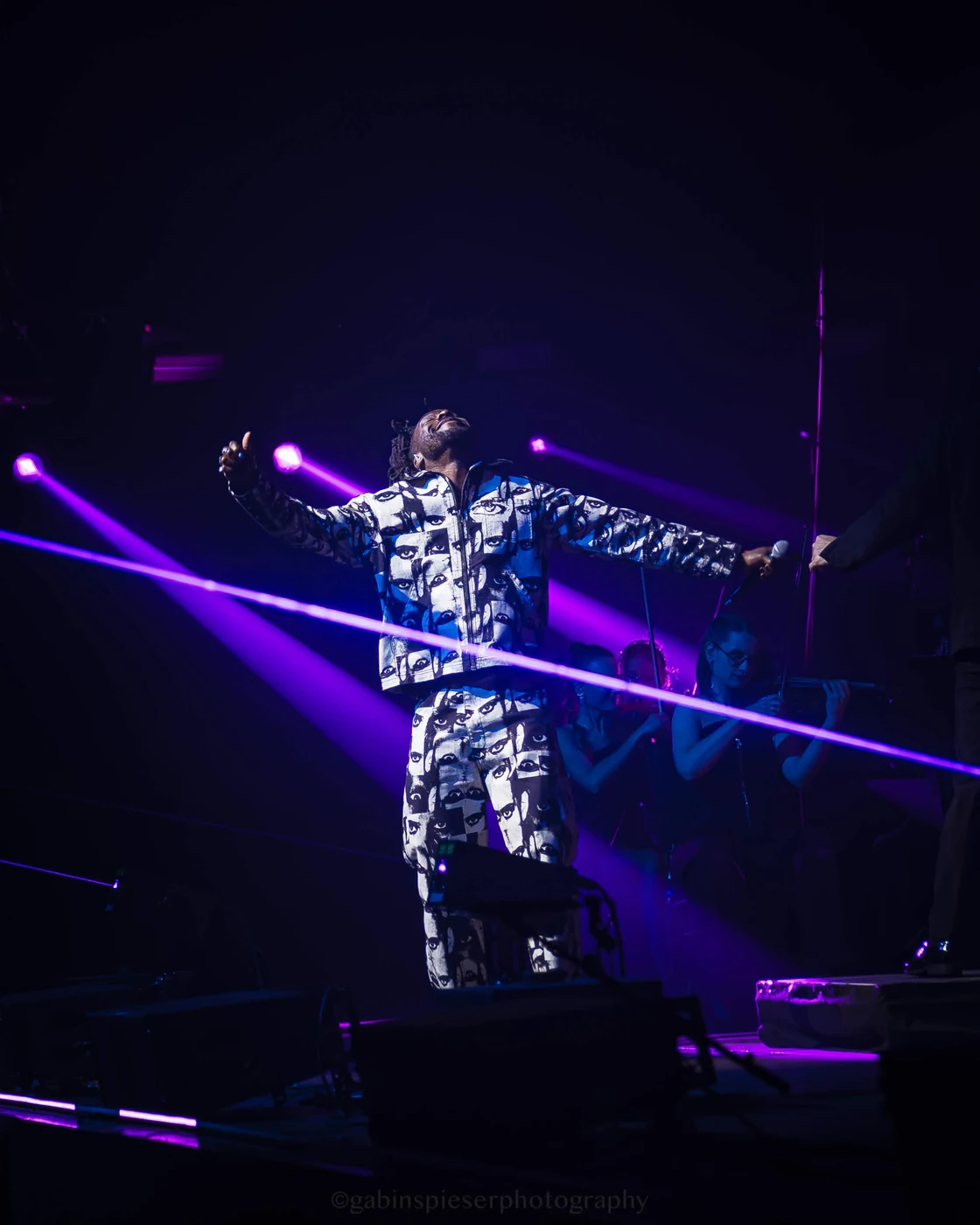 Electro Symphony — Un chanteur en costume à motifs ouvre les bras au milieu de faisceaux violets.