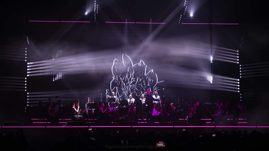 Electro Symphony — Un orchestre et des voix occupent une grande scène traversée de lumières blanches et violettes.