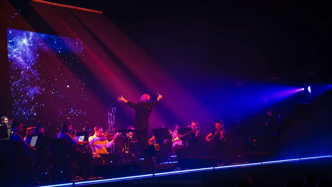 Electro Symphony — Vu de dos, un chef d’orchestre dirige les cordes sous des lumières bleues et roses.