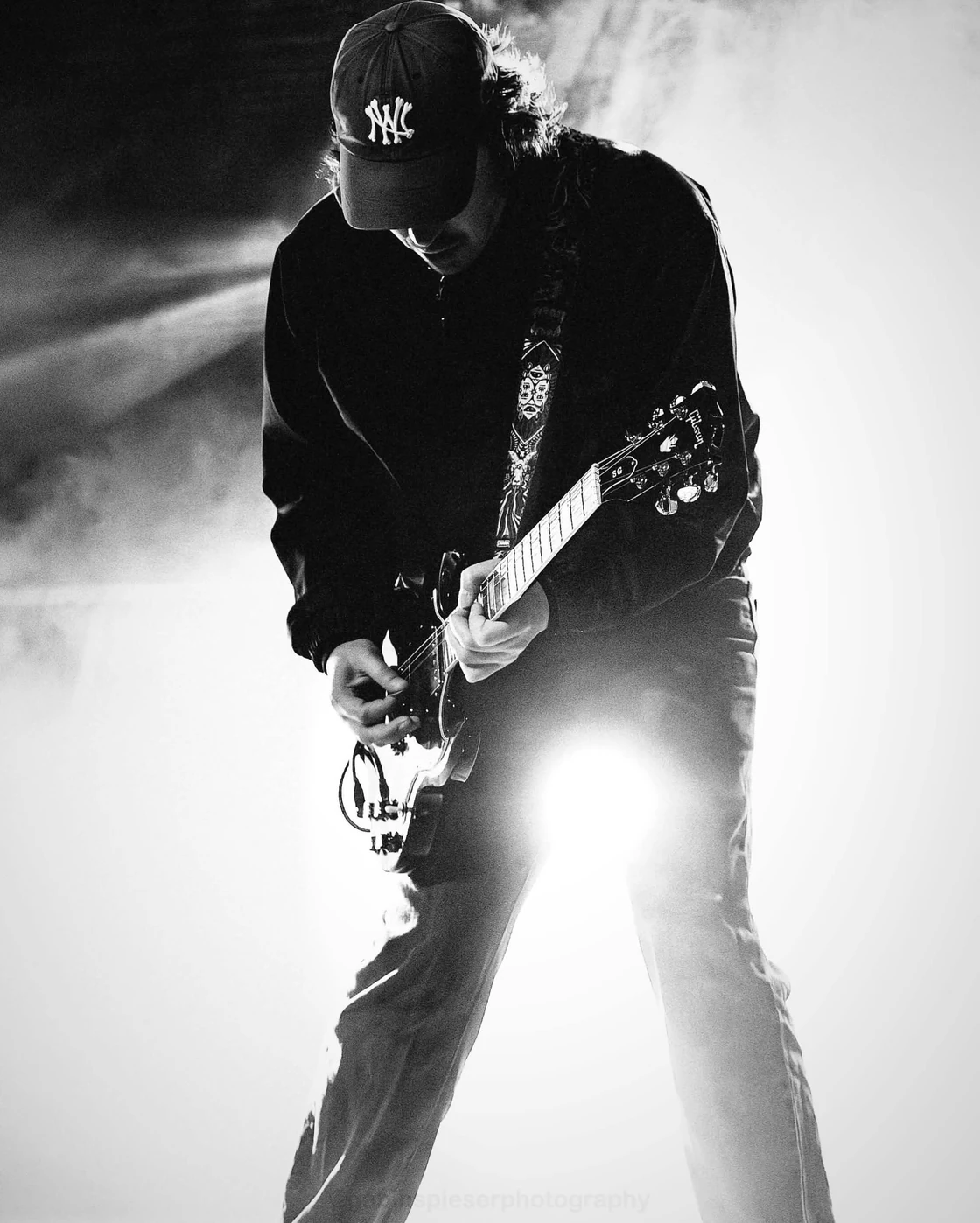 Guitariste de RORI — En noir et blanc, un guitariste en casquette joue au milieu d’une lumière blanche intense. Moissons Rock · mai 2026.