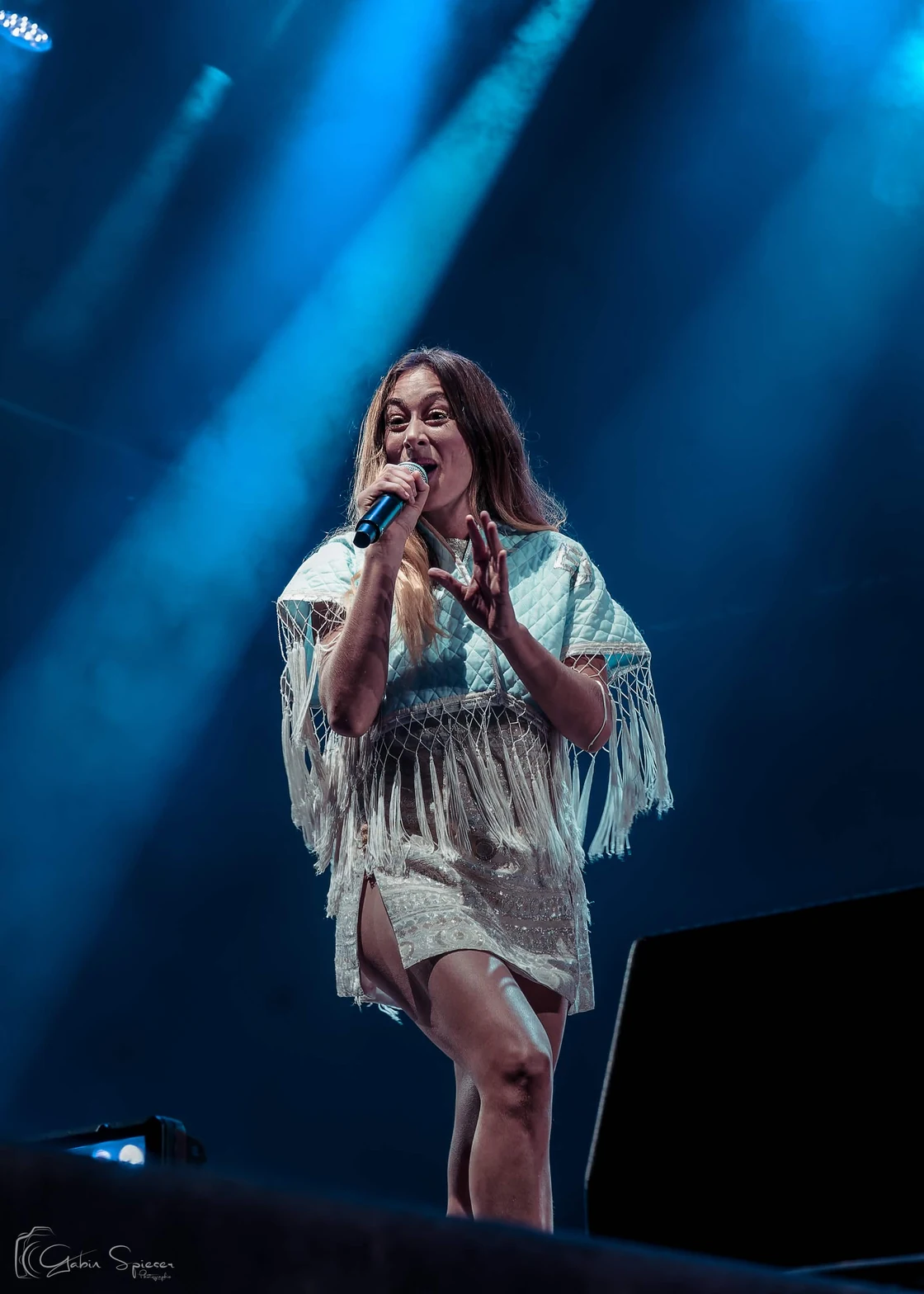 Luiza — Une chanteuse en tenue blanche à franges chante sous des faisceaux de lumière bleue.