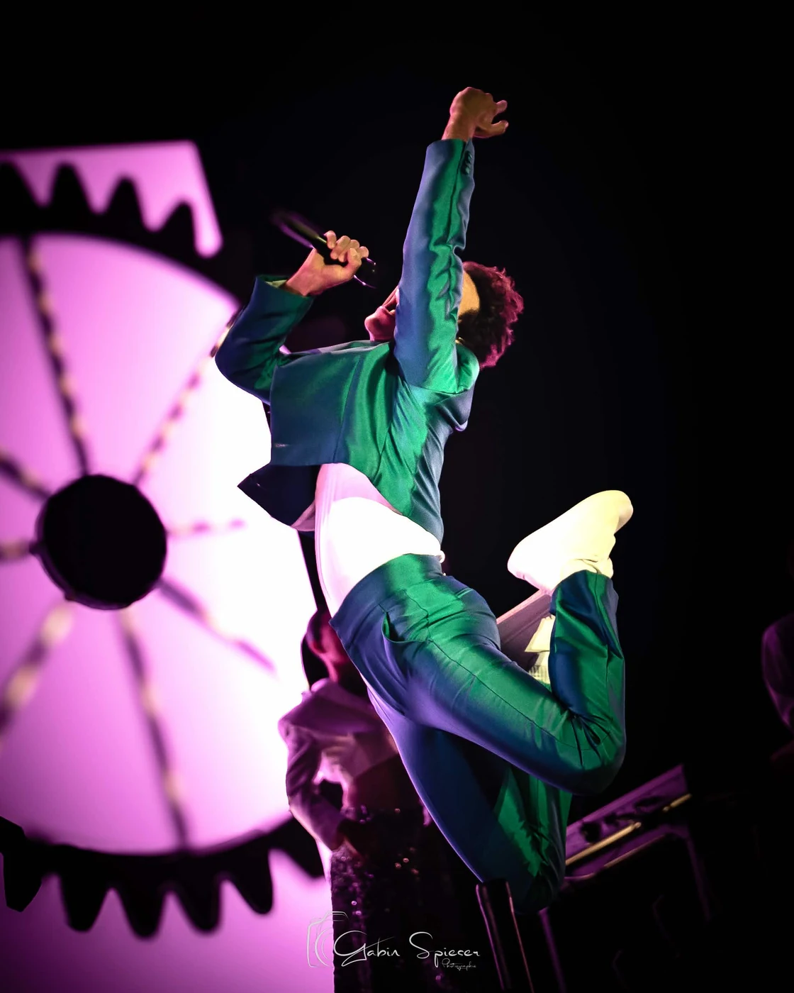 MIKA — Un chanteur en costume vert bondit, micro en main, devant un décor rose lumineux.