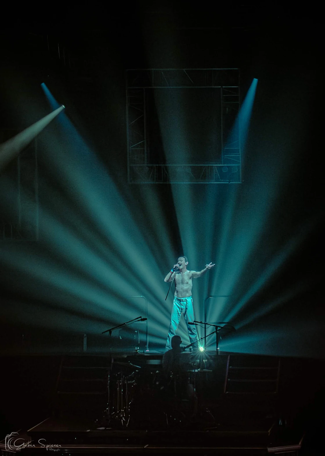 One Night of Queen — Un chanteur au centre d’une scène sombre ouvre un bras devant des faisceaux turquoise.