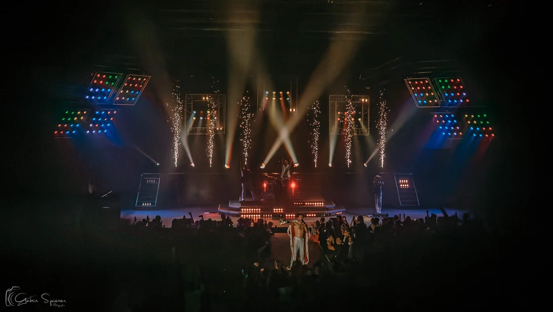 One Night of Queen — Une scène éclairée de faisceaux dorés et d’étincelles, avec un artiste en cape au milieu du public.