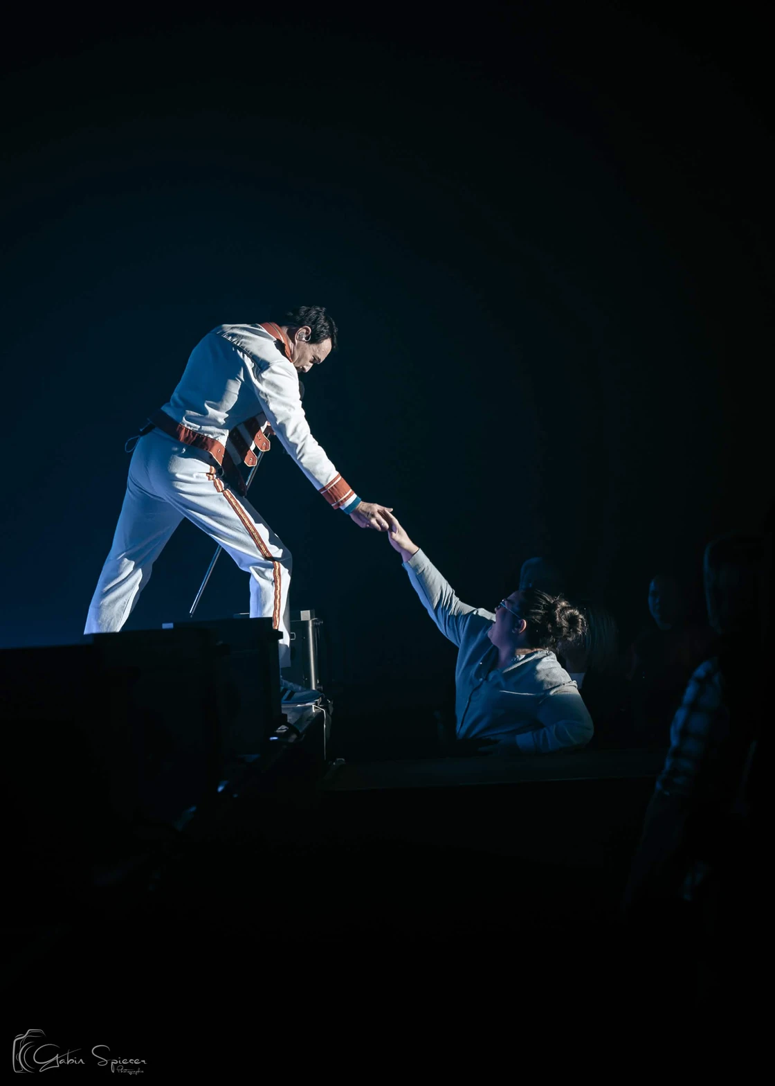 One Night of Queen — Un artiste en costume blanc se penche depuis la scène pour prendre la main d’une personne dans le public.