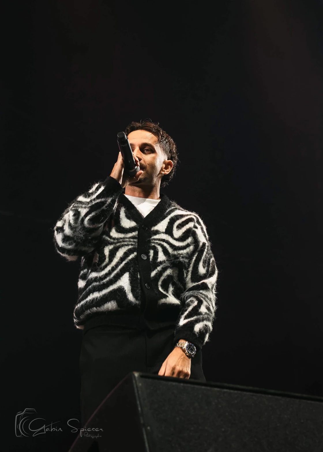 RIDSA — Un chanteur en cardigan noir et blanc au micro, sur une scène sombre.