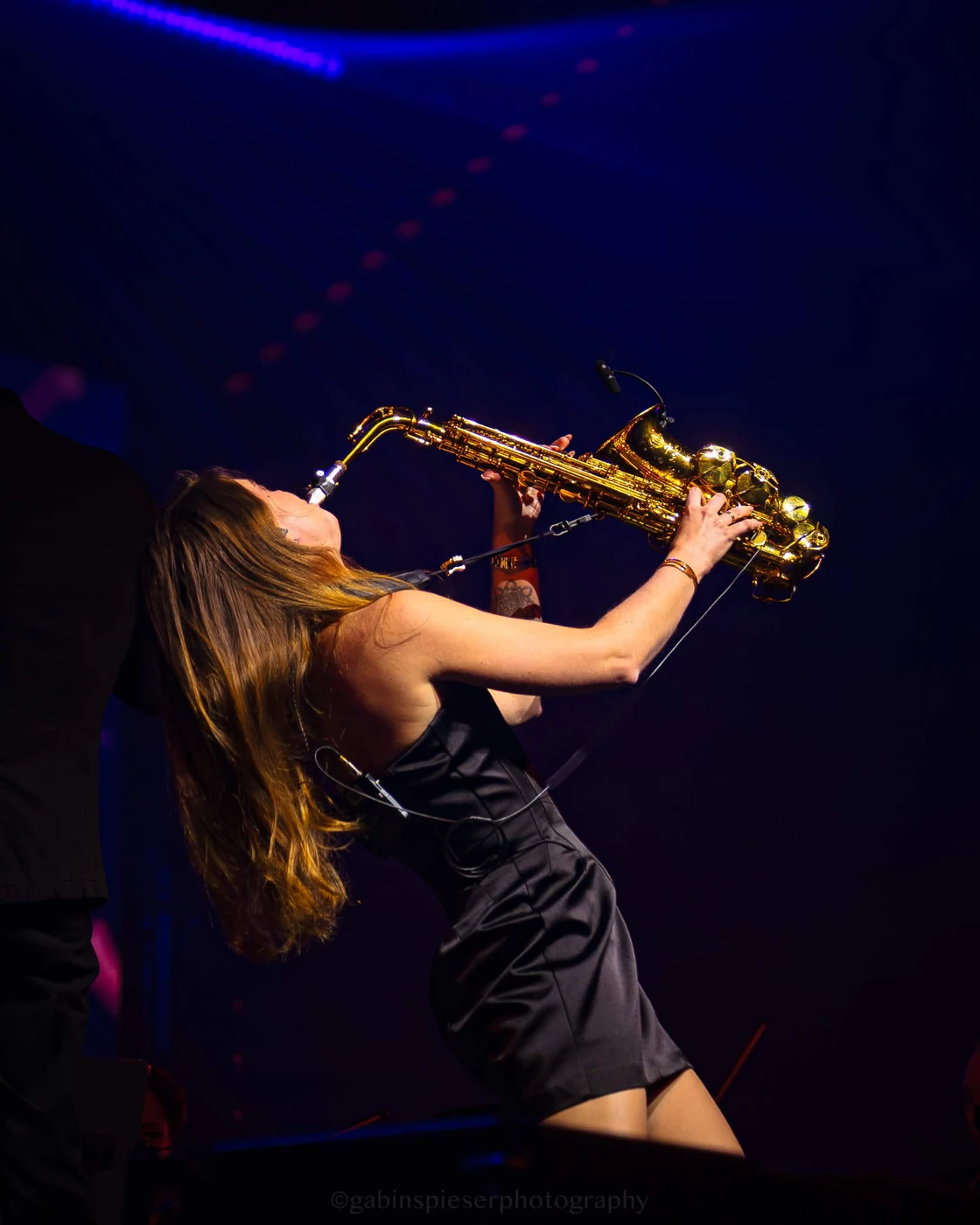 Saxophoniste · Electro Symphony — Une saxophoniste se penche en arrière, son instrument doré levé vers les lumières bleues. Reims Arena · 12 avril 2026.