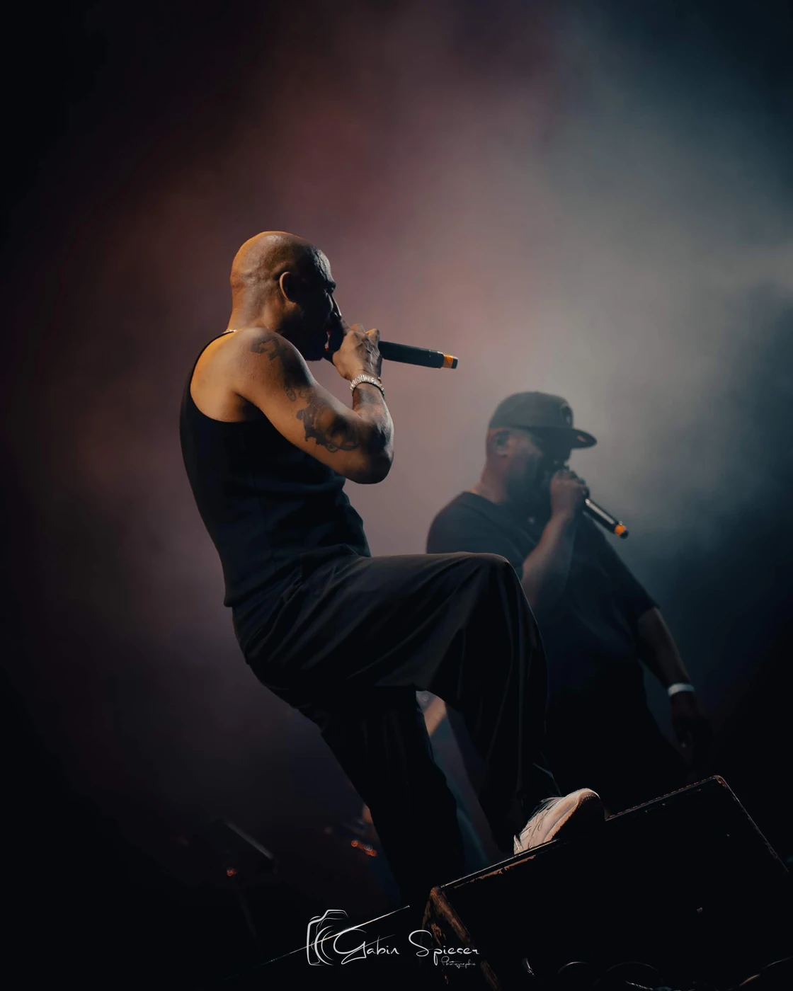 Stomy Bugsy — Deux chanteurs dans la fumée, au premier plan un artiste pose le pied sur un retour de scène.